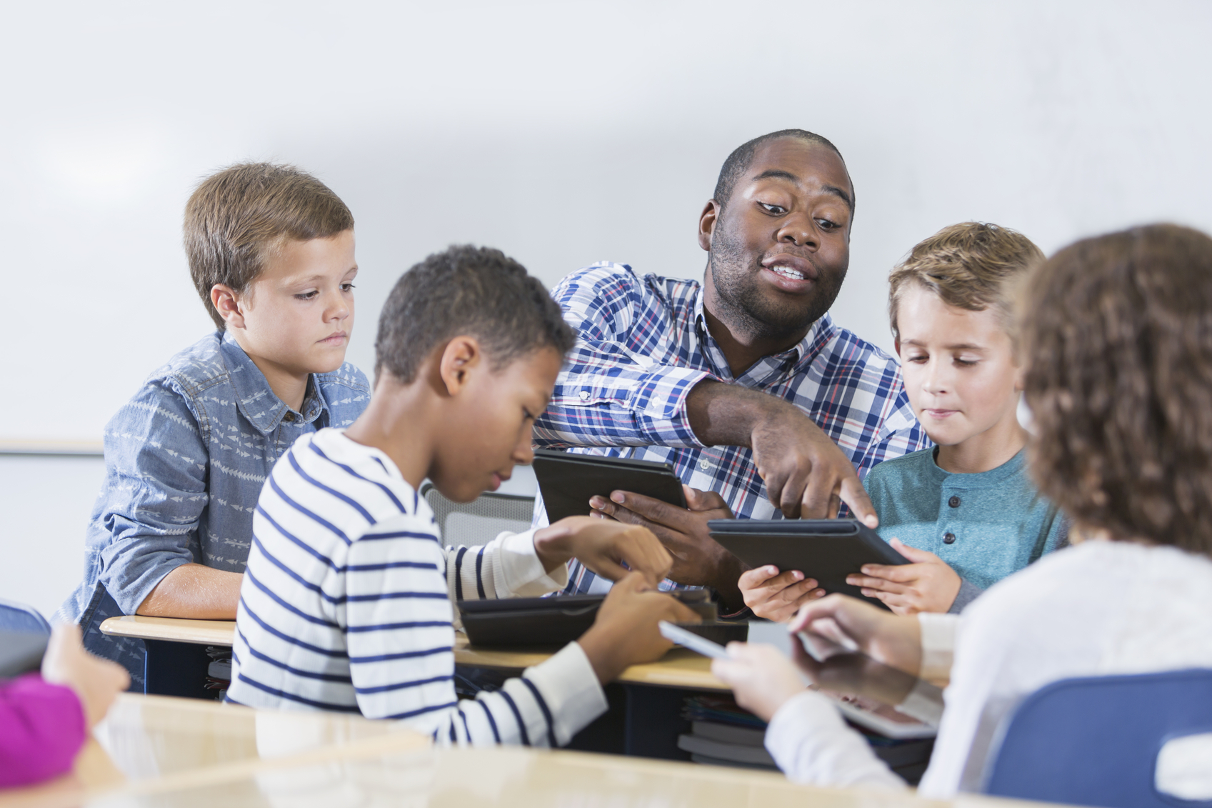A young, African American teacher in class with a group of elementary school students, each with a digital tablet. They are sitting around a table, looking down at the screens, and the man is pointing to the tablet of the boy sitting beside him.