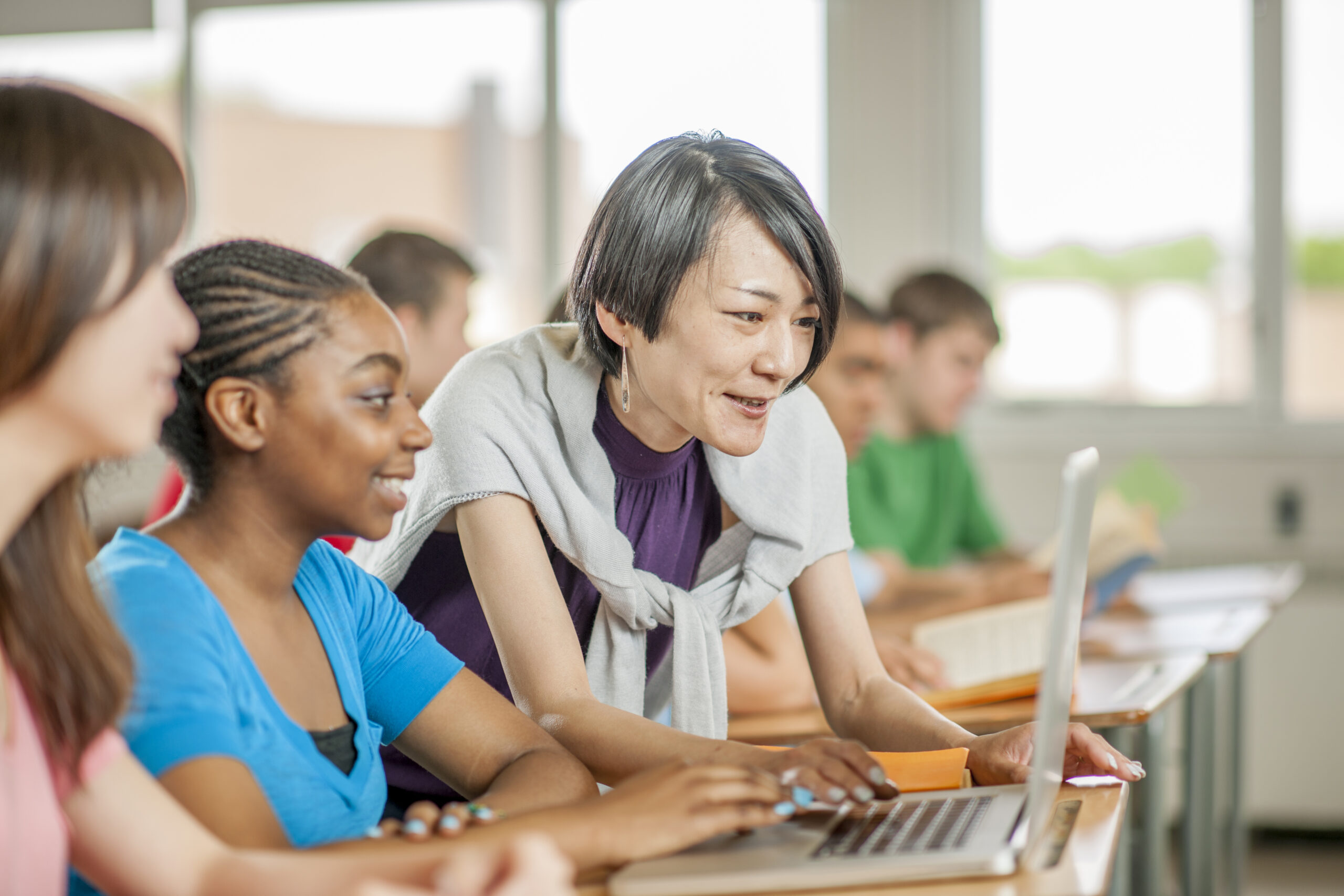 A multi-ethnic group of high school age students are sitting at their desks and are using a laptop to do internet research on an upcoming project. A teacher is helping them look for information.