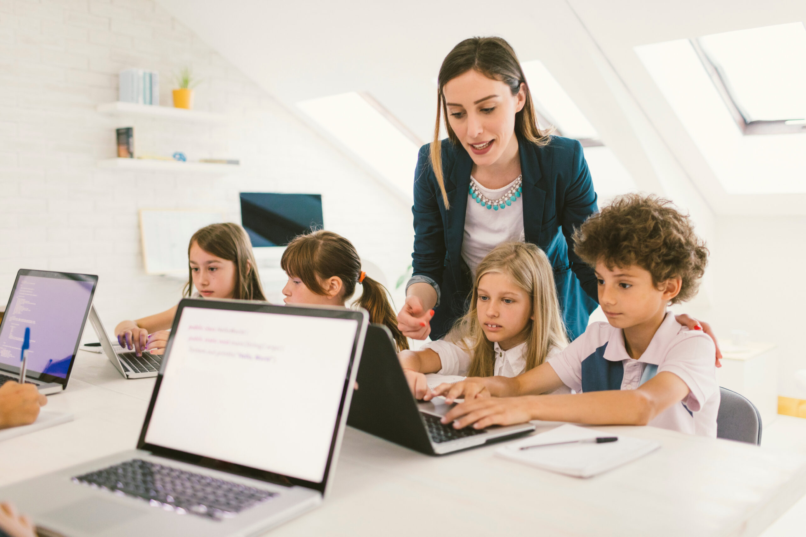 Group of children at coding class. They are sitting by the desk and using laptops. Their female teacher standing beside them and helping with their assignment.